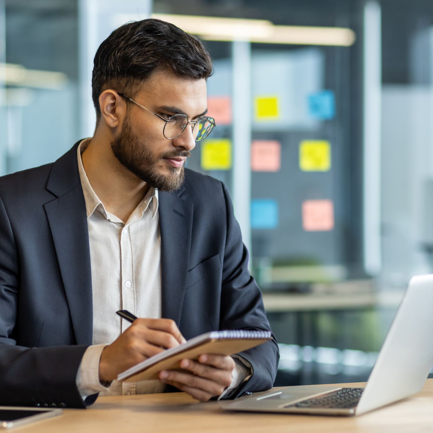 Berater im Anzug sitzt am Schreibtisch, macht Notizen in ein Heft und arbeitet an einem Laptop im Büro.