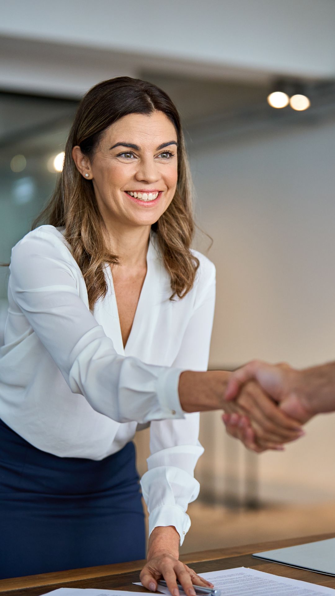 Frau in weißer Bluse reicht bei einem Bewerbungsgespräch die Hand über einen Tisch zum Handschlag.
