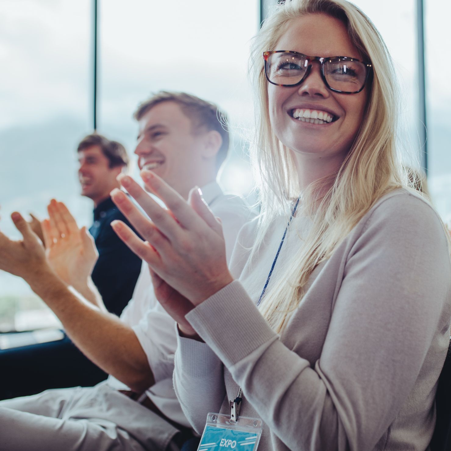 Teilnehmende applaudieren in einem hellen Raum; im Vordergrund eine lächelnde Frau mit Brille.