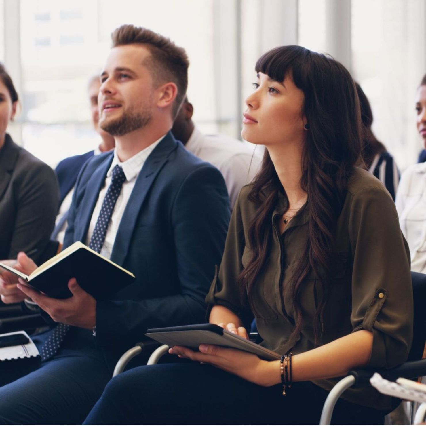 Mehrere Teilnehmende sitzen in einem Seminarraum, hören aufmerksam zu und halten Notizblöcke bzw. Tablets in der Hand.