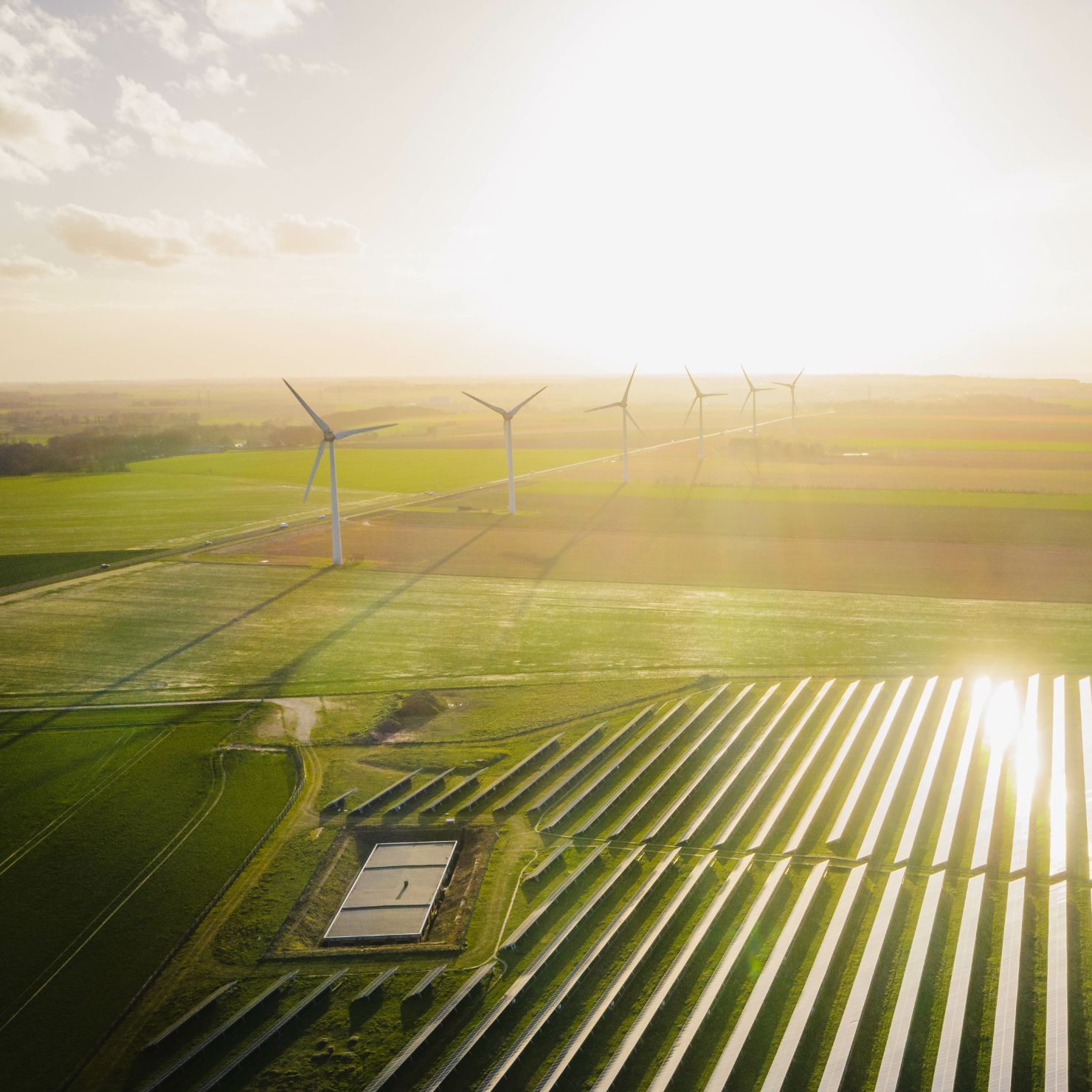 Luftaufnahme eines Solarparks mit Solarmodulen und mehreren Windkraftanlagen in einer ländlichen Landschaft bei Sonnenlicht.